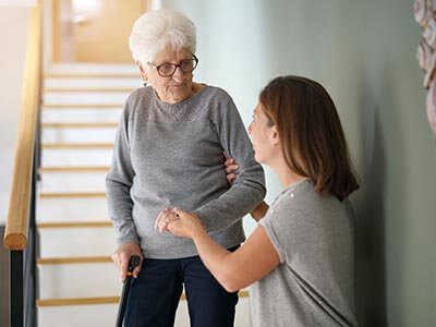 A caring woman assisting an elderly lady up the stairs, providing support and ensuring her safety.